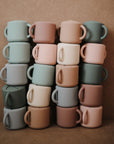 Four vertical stacks of mushie Snack Cups in pastel green, pink, beige, blue, and tan are arranged neatly against a brown background. The small cups resemble a toddler snack container with their soft colors.