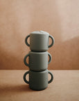 Three gray mushie Snack Cups and toddler cups with handles, made of silicone and featuring a spill-proof design, are stacked vertically against a plain brown, gently lit background.