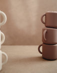 Three brown mugs on the right and three white mugs on the left sit atop a neutral surface with a beige background, creating a calm setting perfect for showcasing mushie's spill-proof Snack Cup for toddlers.