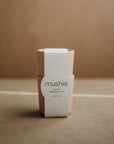 A pair of light pink, non-toxic mushie Dinnerware Cups, dishwasher safe and made in Denmark, are stacked with a white brand band against a brown background.
