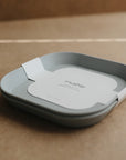 A light blue Square Dinnerware Plate by mushie with rounded edges rests on a beige surface. The BPA-free dish features a white paper label in the center displaying the mushie brand name.
