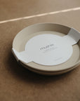 Two beige mushie Round Dinnerware Plates are stacked with a white “mushie” band on a brown textured surface. Made from non-toxic plastic, these plates are dishwasher safe and ideal for everyday use.