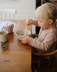 A young blonde child in a pink long-sleeve top sits at a wooden table, eating from a mushie Round Dinnerware Bowl with a spoon. A cup, teapot, and pacifier are on the table as the child focuses on their meal.