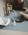 A child’s hands reach toward a running faucet at a modern sink, with an adult nearby. On the matte countertop sits a mushie Finger Toothbrush, along with jewelry in a white shell-shaped dish and a small white object.