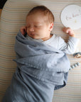 A newborn sleeps peacefully in a light blue mushie Organic Cotton Muslin Swaddle Blanket on a striped mattress, with a round “I’m here!” sun sign in their left hand and a pacifier nearby.