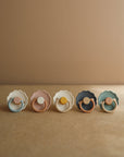 Five FRIGG Daisy Natural Rubber Pacifier 2-Packs in white, pink, brown, dark blue, and green are lined up on a brown surface against a beige background. Each features a daisy-shaped shield and natural rubber latex nipple.