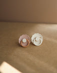 The FRIGG Daisy Natural Rubber Pacifier 2-Pack by FRIGG features pink and white daisy-shaped pacifiers with scalloped edges and natural rubber latex nipples, displayed side by side on a smooth brown surface under soft lighting.