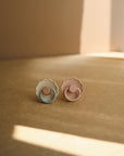 Two FRIGG Daisy Natural Rubber Pacifiers—one blue/beige, one pink/beige—from the FRIGG Daisy 2-Pack rest on a brown surface in soft sunlight and gentle shadows.