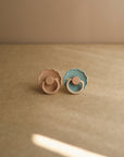 The FRIGG Daisy Natural Rubber Pacifier 2-Pack by FRIGG features a light beige and a pastel blue pacifier with natural rubber latex nipples, shown upright on a tan surface with a soft beige background under gentle lighting.