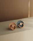 The FRIGG Daisy Natural Rubber Pacifier 2-Pack features two scallop-edged, daisy-shaped pacifiers—one in muted peach and one in dark gray—standing upright on a softly lit beige surface with a warm, neutral backdrop.