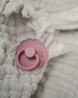 The FRIGG Daisy Natural Rubber Pacifier 2-Pack, featuring a daisy-shaped design and natural rubber nipple, is shown resting on textured off-white fringed fabric.