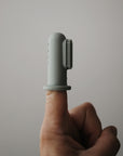 A hand with an extended index finger wears the mushie Finger Toothbrush, a silicone brush with short bristles ideal for gentle baby teeth cleaning, set against a plain gray background.