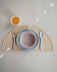 The mushie Round Dinnerware Bowl set includes a pastel yellow cup, pink fork and spoon, and BPA-free baby bowls on a pink plate, all arranged on a beige half-circle placemat against a light background.