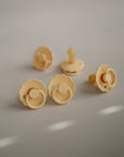 Five FRIGG Daisy Natural Rubber Pacifier 2-Packs, featuring yellow daisy-shaped designs and natural rubber nipples, are arranged on a light grey surface with soft lighting casting gentle shadows and highlights.