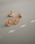 Two pastel pink FRIGG Daisy Natural Rubber Pacifiers from the FRIGG Daisy Natural Rubber Pacifier 2-Pack are in the foreground and one is in the background on a light gray surface, softly lit to create gentle shadows and highlights.