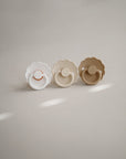 Two FRIGG Daisy Natural Rubber Pacifiers from the 2-Pack, featuring scalloped edges and natural rubber latex nipples, are displayed upright on a beige surface in white and taupe hues under soft natural lighting.