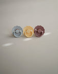 FRIGG Daisy Natural Rubber Pacifier 2-Pack by FRIGG features pastel blue, yellow, and mauve pacifiers with scalloped edges and a minimal design, each with a natural rubber latex nipple. Photographed upright on a softly lit surface.