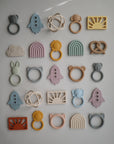 A colorful grid of mushie silicone baby teethers, including the Pretzel Teether and other non-toxic shapes like rockets, rainbows, animals, and rings, displayed on a white background.