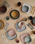 Mushie Square Dinnerware Plates, BPA-free and pastel-hued in soft yellow, pink, gray, and brown tones, are pictured neatly arranged with matching bowls, cups, forks, and spoons on a beige surface.