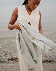 A woman stands on the beach holding the mushie Organic Cotton Muslin Swaddle Blanket with beaded pacifier clips. She wears a cream sleeveless top and pants, with the shoreline and water in the background.