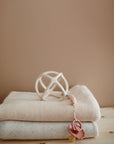 Two folded beige baby blankets rest on a wooden surface, topped with a white geometric rattle and mushie's Silicone Pacifier Clip | Hera. A pacifier is attached to the clip. The plain background is a soft brown wall.