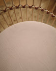 Close-up of the mushie Mini Muslin Crib Sheet in light pink, made from 100% cotton with a subtle texture, displayed on a cushioned seat with curved light-colored rods and brown bindings.