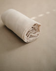 The mushie Extra Soft Bassinet Muslin Crib Sheet is neatly rolled on a smooth, light beige surface with soft lighting, creating a calm and minimalistic vibe—perfect for breathable baby bedding.