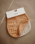 The mushie Organic Cotton Muslin Burp Cloth 2-Pack features soft, organic cotton burp cloths in beige and light brown, shown overlapping on a neutral background with the mushie label attached at the top.