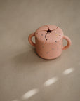 The mushie Snack Cup is a small, pink speckled silicone cup with two handles and a soft, spill-proof slotted lid—perfect for toddlers. It sits on a beige surface with light spots.