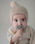 A baby in a beige knit hat and cream outfit holds the FRIGG Daisy Natural Rubber Pacifier to their mouth, looking up wide-eyed against a simple background. Brand: FRIGG, Product: Daisy Natural Rubber Pacifier 2-Pack.