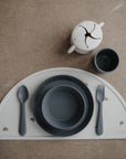 The mushie Round Dinnerware Plates set features dishwasher-safe minimalist children’s tableware—a grey bowl, plate, spoon, and fork on a rainbow semicircular mat—plus a matching grey cup and cream snack cup displayed on a brown surface.
