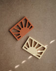 Two mushie Sun Teethers in terracotta and beige, both featuring sunburst designs, rest on a brown textured surface with light streaks. These non-toxic teethers provide style and safety for your little one.