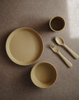 A set of mustard yellow mushie Round Dinnerware Plates in non-toxic, dishwasher-safe silicone on a brown surface, with two bowls, cup, fork, and spoon. The plate and large bowl are embossed with “mushie.”.