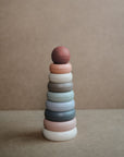 The mushie Stacking Rings Toy is a non-toxic baby toy with pastel rings sized to stack and a reddish sphere on top, shown on a neutral brown background.