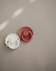 Two FRIGG Daisy Natural Rubber Pacifiers—one cream and one rust—with scalloped edges and natural rubber nipples rest on a neutral beige surface in soft light. Available as a 2-pack from FRIGG.
