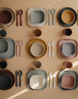 A flat lay of colorful, neatly arranged mushie Round Dinnerware Plates—non-toxic, dishwasher safe, and in muted earth tones—displayed on a peach background with light and shadow patterns.