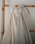 Two mushie Organic Cotton Muslin Swaddle Blankets—one cream with florals, one light blue with white stripes—are tied to a wooden ladder set against a plain white wall.