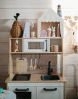 The mushie Stacking Rings Toy is displayed with a pretend sink, stove, microwave, pastel accessories, wooden utensils, and non-toxic baskets against a light wood-paneled wall in soft natural light.