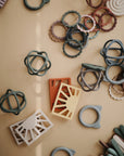 The mushie Sun Teether and other assorted silicone baby toys in soft, muted colors are scattered on a beige surface, featuring non-toxic materials and safe rounded or geometric designs for playful teething and stacking.