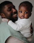 A smiling man holds a baby in white, with a mushie Organic Cotton Muslin Burp Cloth from the 2-Pack draped over his shoulder. The softly blurred background keeps the focus on them.