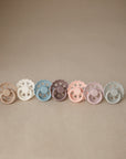 Seven FRIGG Andersen Fairytale Silicone Pacifier 2-Packs in muted shades (tan, cream, gray, brown, mauve, pink, blue) are displayed on a beige surface with matching background—each pacifier is thoughtfully made in Denmark.