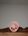 The FRIGG Andersen Fairytale Silicone Pacifier 2-Pack by FRIGG, featuring heart-shaped cutouts and made in Denmark, stands upright on a rustic wooden surface against a plain gray background.