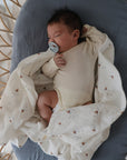 A dark-haired baby sleeps on a blue cushion, wrapped in a mushie Organic Cotton Muslin Swaddle Blanket with subtle patterns, wearing a cream onesie and sucking a pacifier.