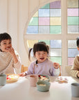 Three young children sit at a table with mushie Square Dinnerware Plates, smiling as they eat snacks. A colorful stained glass window in the background lets in soft light.