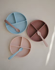 Three round sectioned plates in blue, pink, and mauve are arranged with mushie Silicone Feeding Spoons. Each plate has three compartments for organized meals and comes with a soft baby feeding spoon.