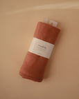 A Ribbed Baby Blanket by Mushie in rust color, made from organic cotton, is rolled up with a white Mushie label and placed on a beige background.