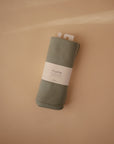 A sage green Ribbed Baby Blanket from Mushie, neatly rolled with a white "mushie" label, rests on a light beige surface.