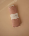 A rolled-up Mushie Ribbed Baby Blanket in peach, made from organic cotton, sits on a beige surface. The breathable blanket features a white Mushie label and two white tags at the top.