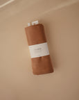A rolled-up Mushie Ribbed Baby Blanket in brown, featuring a white Mushie label, rests on a beige surface.