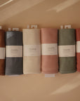Six ribbed Mushie organic cotton baby blankets in earthy tones are lined up side by side. Each breathable blanket features a white Mushie label on a warm beige background.